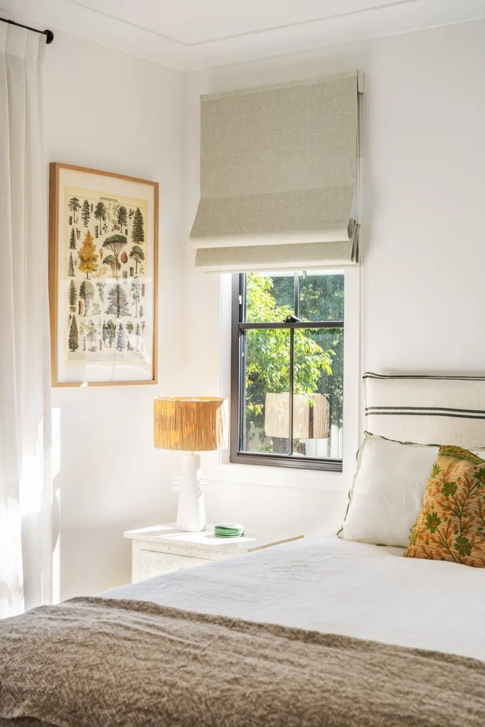 A bedroom with neutral tones, a striped bedhead, botanical art print on the wall and a bedside table with a lamp on top.