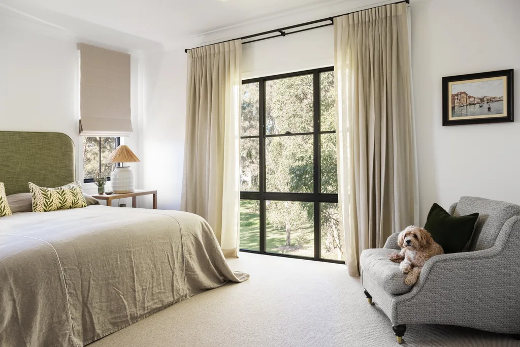 A neutral toned bedroom with a green bedhead, grey armchair in the corner and a dog sitting on it.