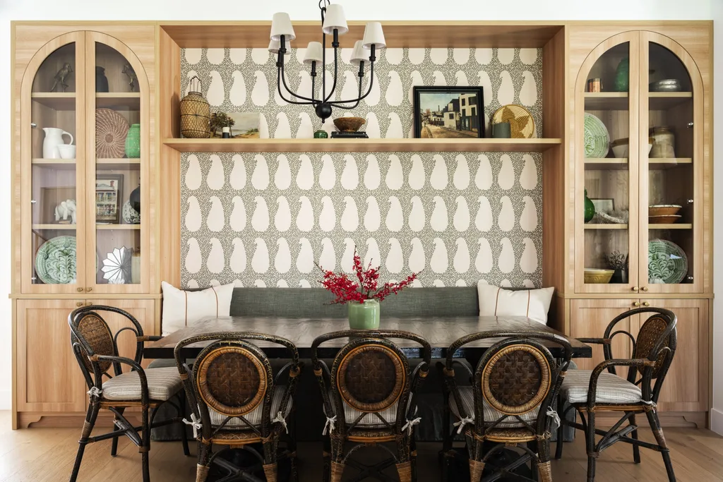 A wall has two timber and glass cabinets with built in banquette seating in between. A dining table is positioned beside this banquette seat, with black and rattan dining chairs on the opposite side.
