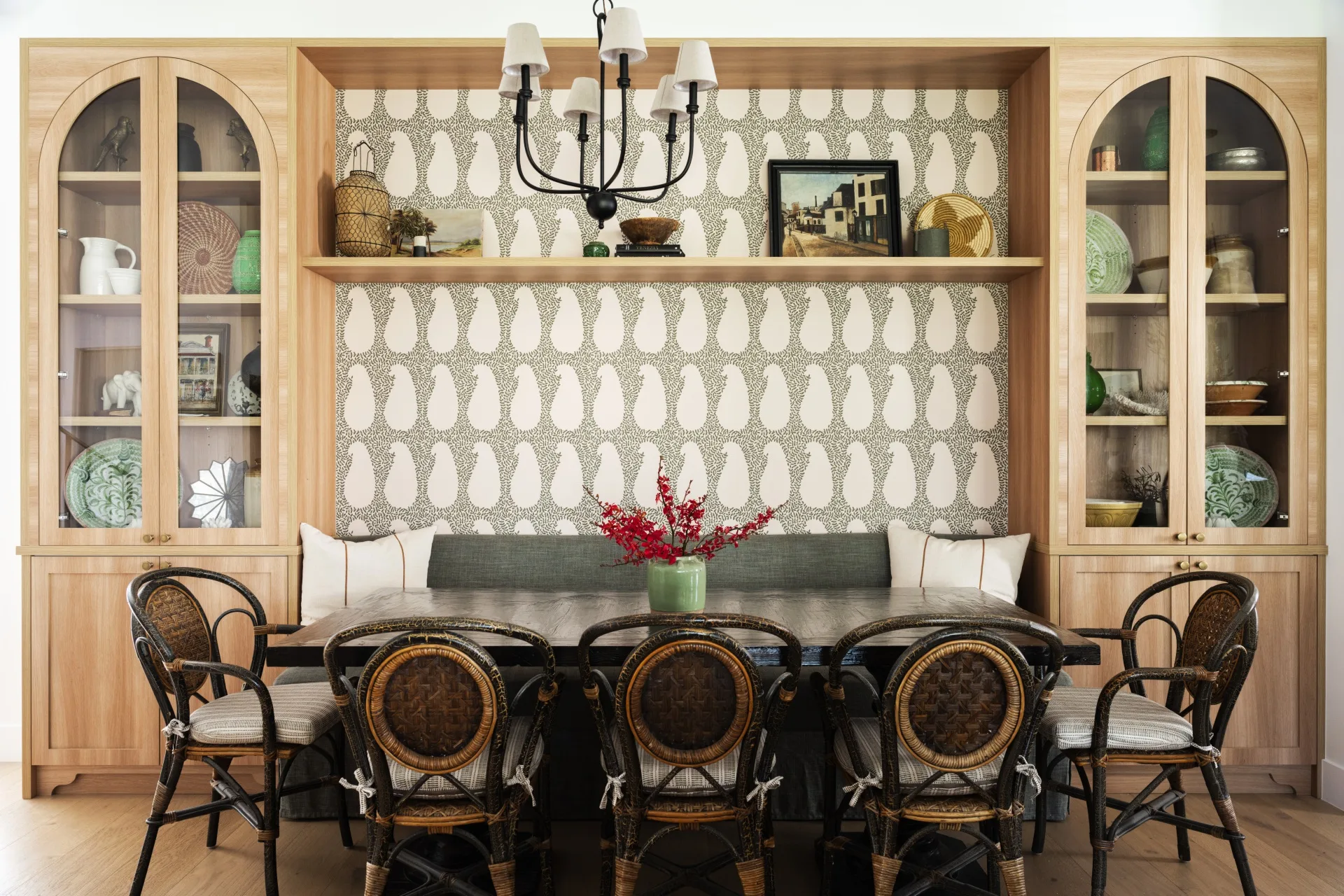 A wall has two timber and glass cabinets with built in banquette seating in between. A dining table is positioned beside this banquette seat, with black and rattan dining chairs on the opposite side.