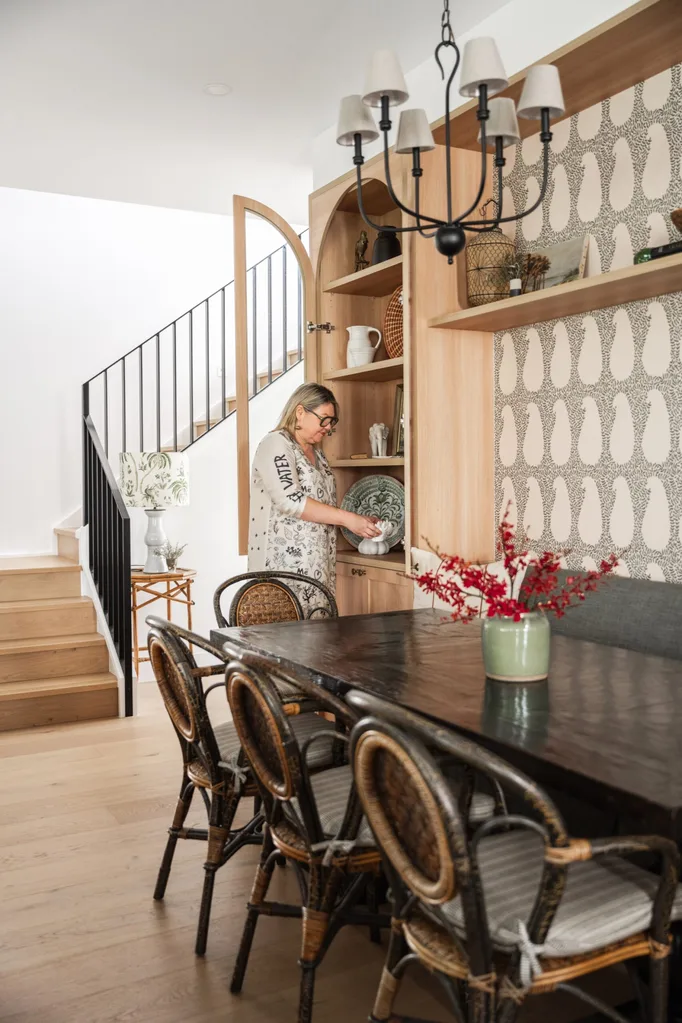 A wall has two timber and glass cabinets with built in banquette seating in between. A dining table is positioned beside this banquette seat, with black and rattan dining chairs on the opposite side. A woman looks at an object in the cabinet.