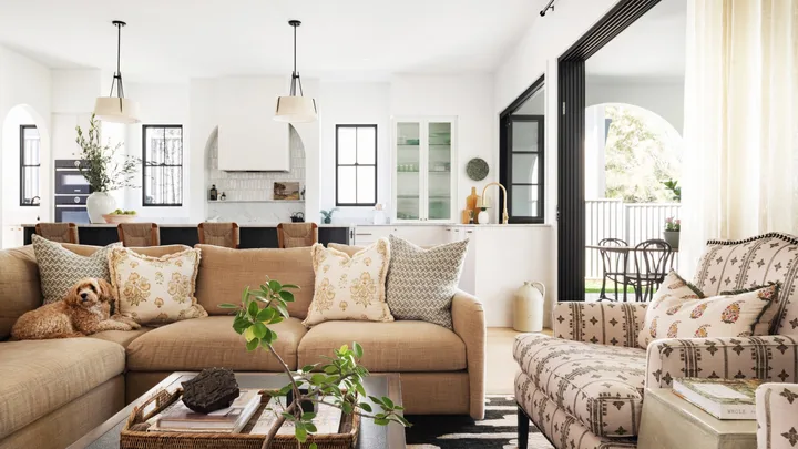 An open plan kitchen and living room which opens out to an alfresco dining and living area. A dog sits on the neutral toned couch. The space is primarily black and white, with warm tones in the living room seats.
