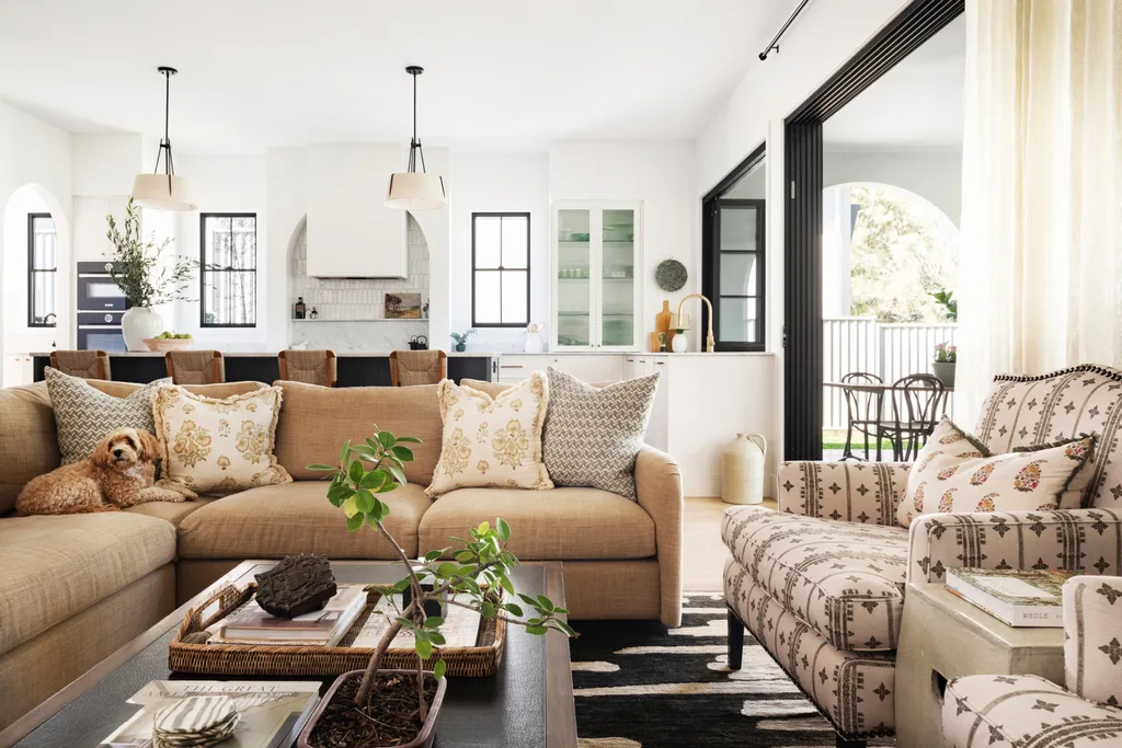 An open plan kitchen and living room which opens out to an alfresco dining and living area. A dog sits on the neutral toned couch. The space is primarily black and white, with warm tones in the living room seats.