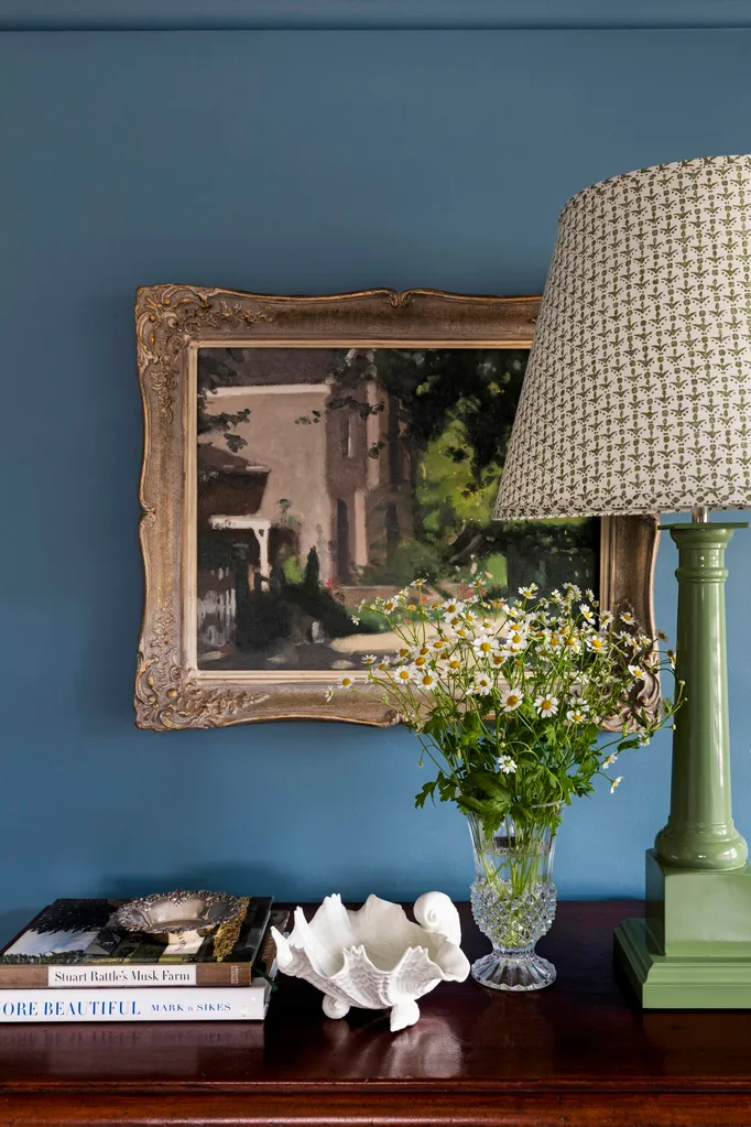 A close up photo of the top of a timber side table, with walls painted blue, a framed painting mounted on the wall and a green lamp base with patterned lampshade. The palette feels perfect for winter decorating and could inspire winter decorating ideas.