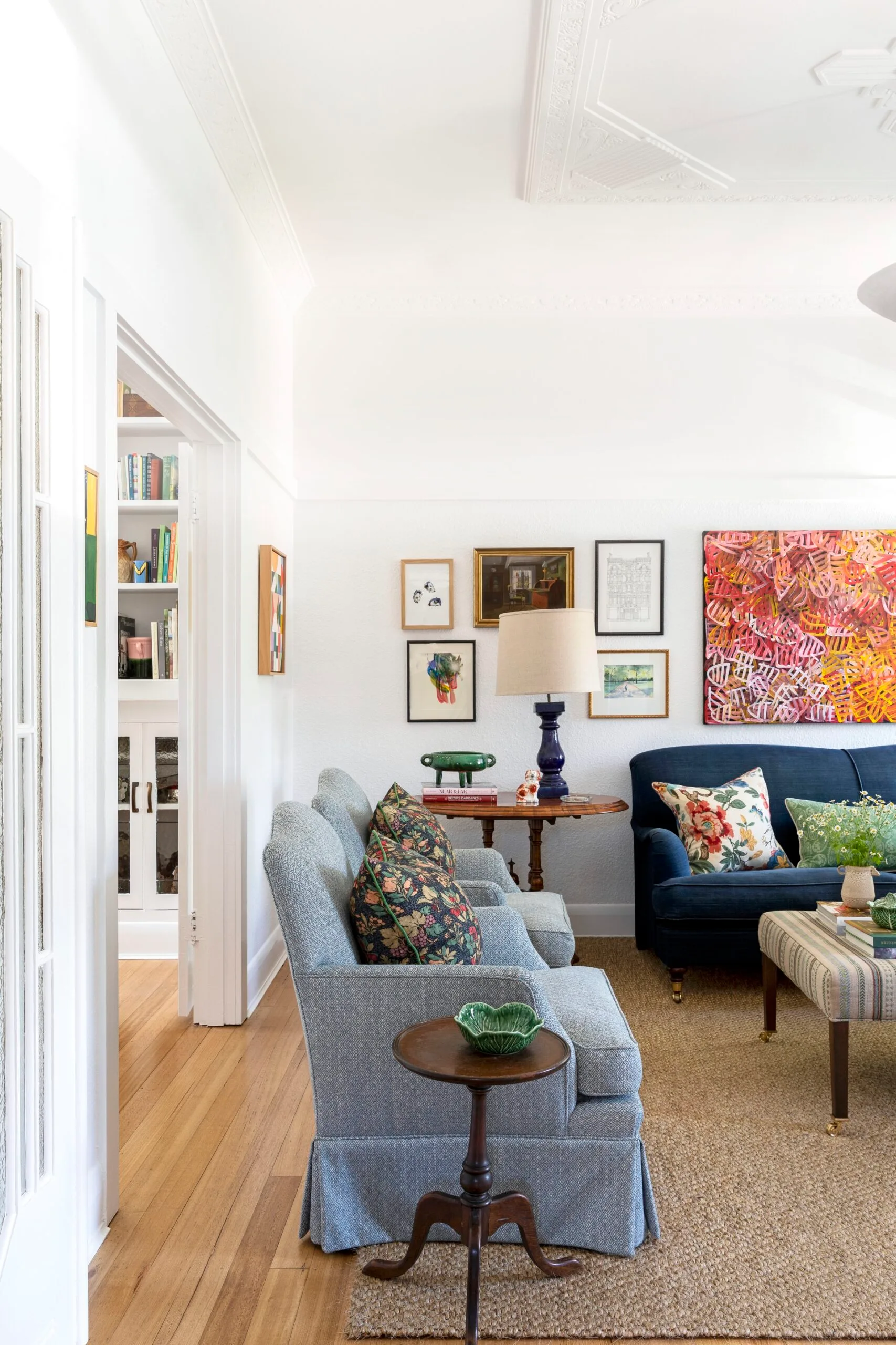 A living room with white walls and ceiling, blue couches on top of a sisal rug and a striped ottoman which doubles as a coffee table. A gallery wall adds colour and a door leads to a dining area. The palette feels perfect for winter decorating and could inspire winter decorating ideas.