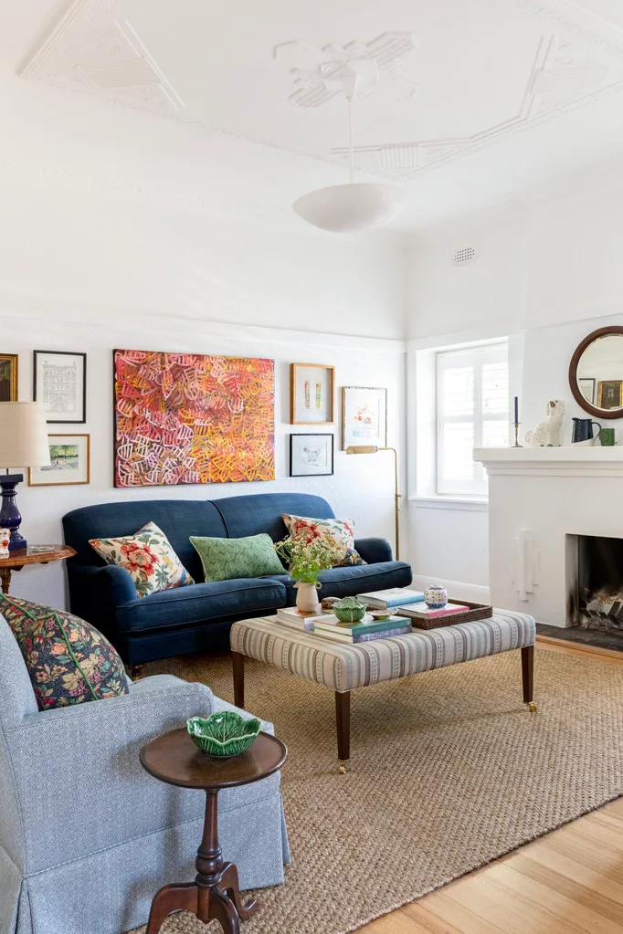 A living room with white walls and ceiling, blue couches on top of a sisal rug and a striped ottoman which doubles as a coffee table. A fireplace has a round mirror above it. The palette feels perfect for winter decorating and could inspire winter decorating ideas.