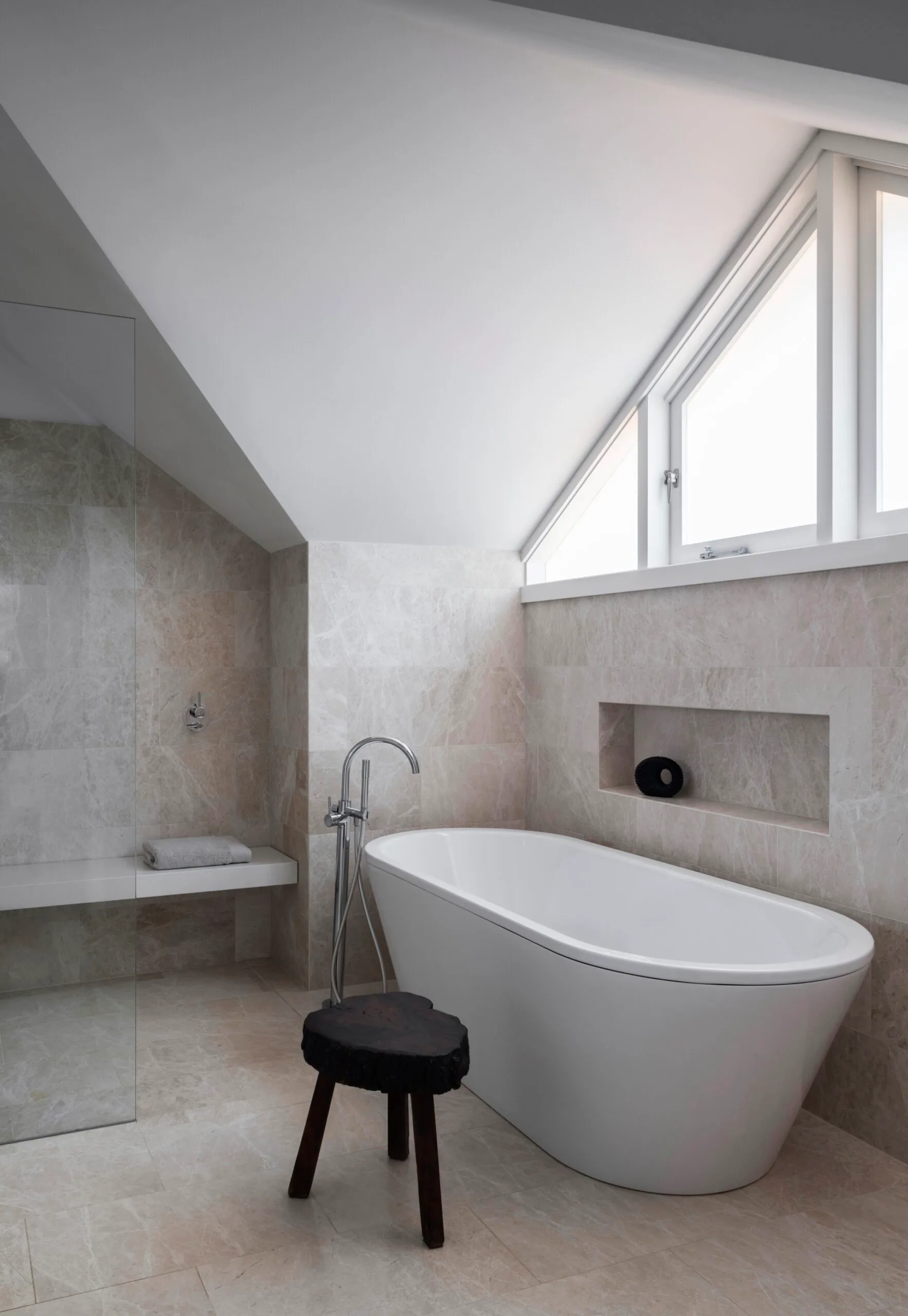 An ensuite bathroom with large marble tiles on the floor and walls, a marble benchtop on the vanity, white paint on the slanted roofline, a shower with a shower bench and a freestanding bath.