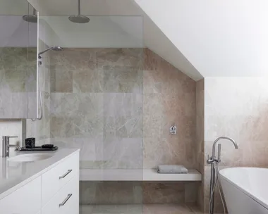 An ensuite bathroom with large marble tiles on the floor and walls, a marble benchtop on the vanity, white paint on the slanted roofline, a shower with a shower bench and a freestanding bath.