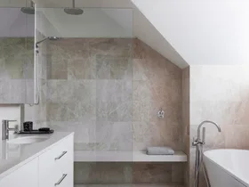 An ensuite bathroom with large marble tiles on the floor and walls, a marble benchtop on the vanity, white paint on the slanted roofline, a shower with a shower bench and a freestanding bath.