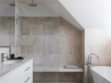 An ensuite bathroom with large marble tiles on the floor and walls, a marble benchtop on the vanity, white paint on the slanted roofline, a shower with a shower bench and a freestanding bath.