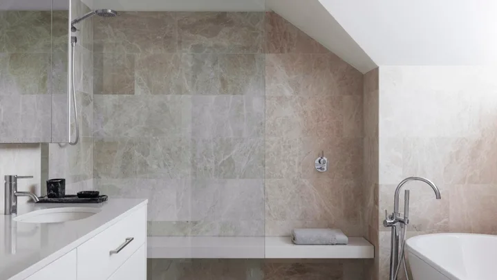 An ensuite bathroom with large marble tiles on the floor and walls, a marble benchtop on the vanity, white paint on the slanted roofline, a shower with a shower bench and a freestanding bath.
