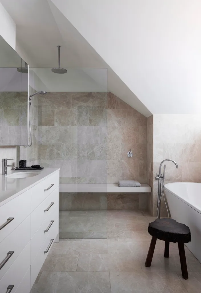 An ensuite bathroom with large marble tiles on the floor and walls, a marble benchtop on the vanity, white paint on the slanted roofline, a shower with a shower bench and a freestanding bath.