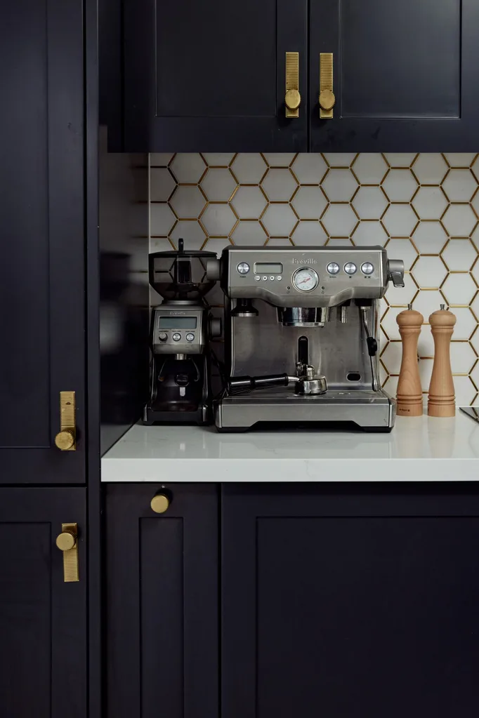 Dark kitchen cabinetry with brass handles. A stainless steel espresso coffee machine is on a white benchtop, in front of white hexagonal tiled splashback.