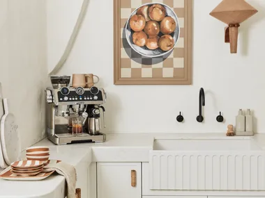 A DeLonghi coffee machine with built in milk steamer and coffee grinder on a white kitchen bench with white cabinetry and white walls.