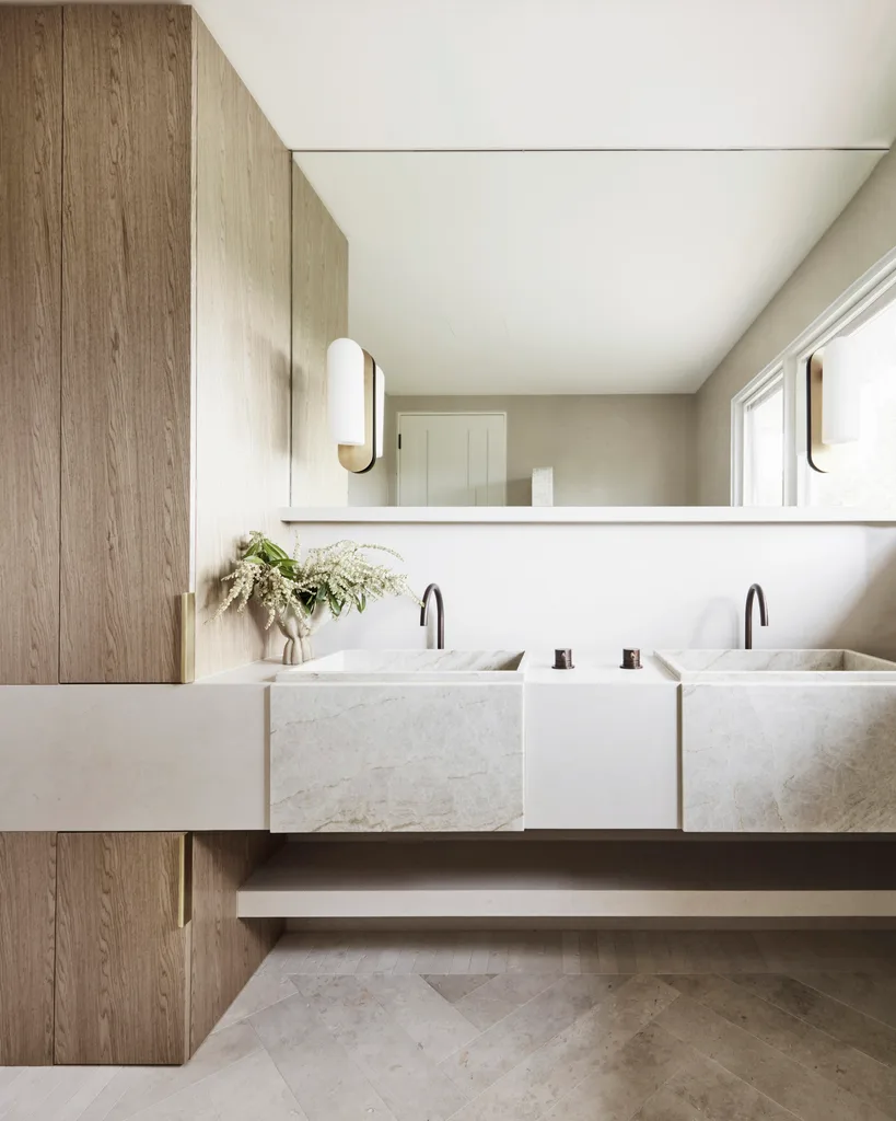 Two basins are side by side in a vanity, beneath a mirror and two wall lights. Earth tones fit in to the earthy colour palette, with gooseneck taps in weathered brass organic and honed quartzite for the basins. The floor tiles are raw-sawn limestone laid in a herringbone pattern.