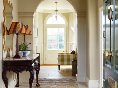 A hallway entrance to a home, with tiled flooring and a rug placed atop. This liminal space has a console table with two lamps on top and a gold-framed mirror on the wall.