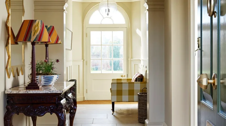 A hallway entrance to a home, with tiled flooring and a rug placed atop. This liminal space has a console table with two lamps on top and a gold-framed mirror on the wall.
