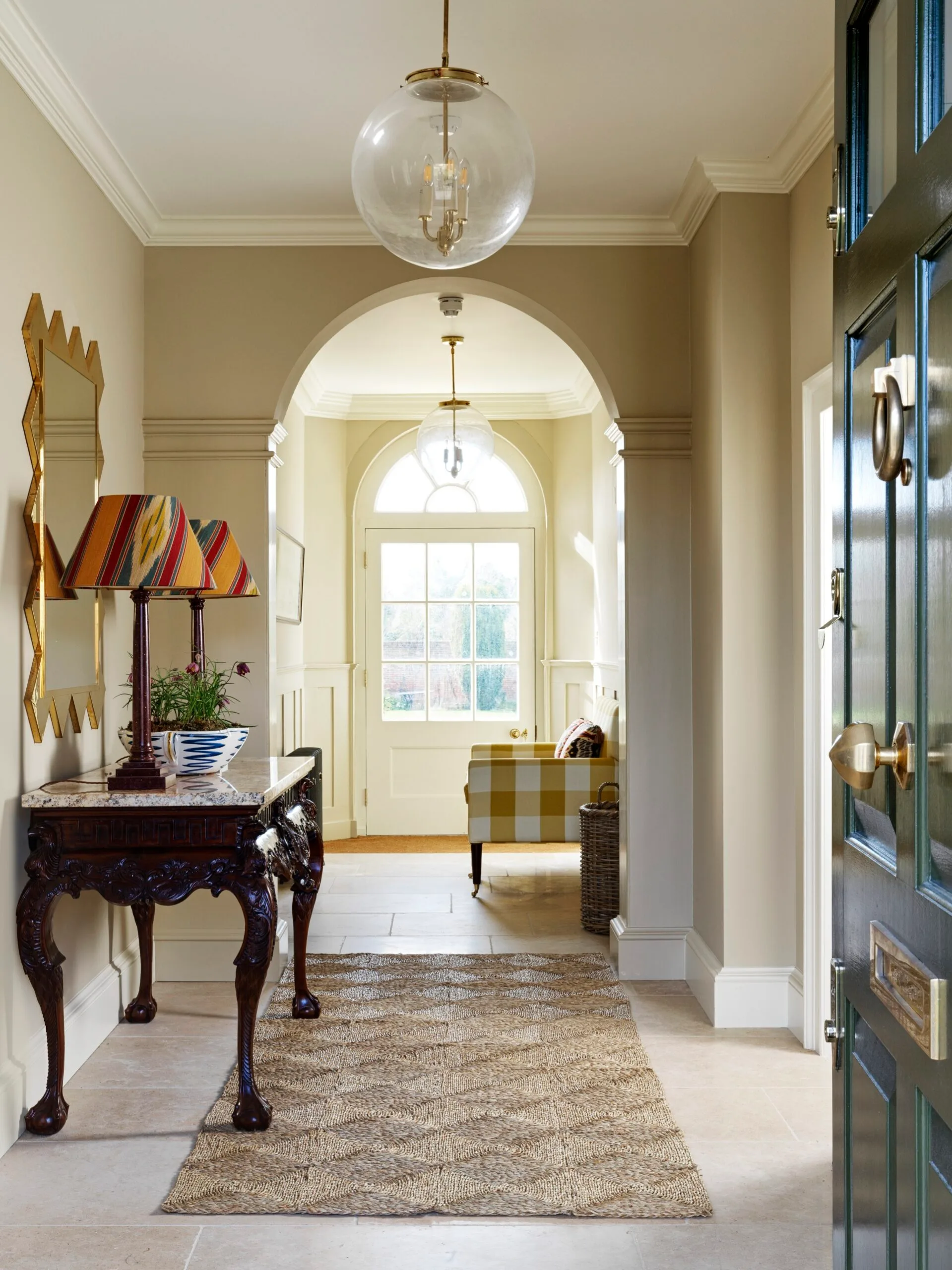 A hallway entrance to a home, with tiled flooring and a rug placed atop. This liminal space has a console table with two lamps on top and a gold-framed mirror on the wall.