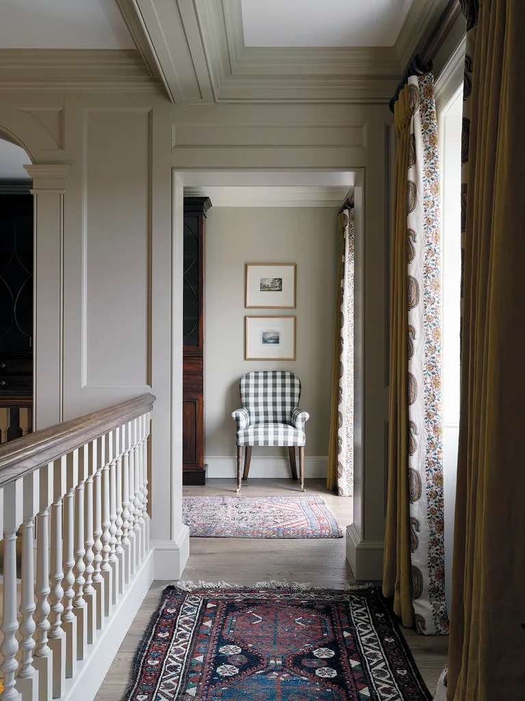 A liminal hallway on the second floor of a home, with coffered ceilings, traditional patterned rugs, heavy curtains and a checkered armchair.