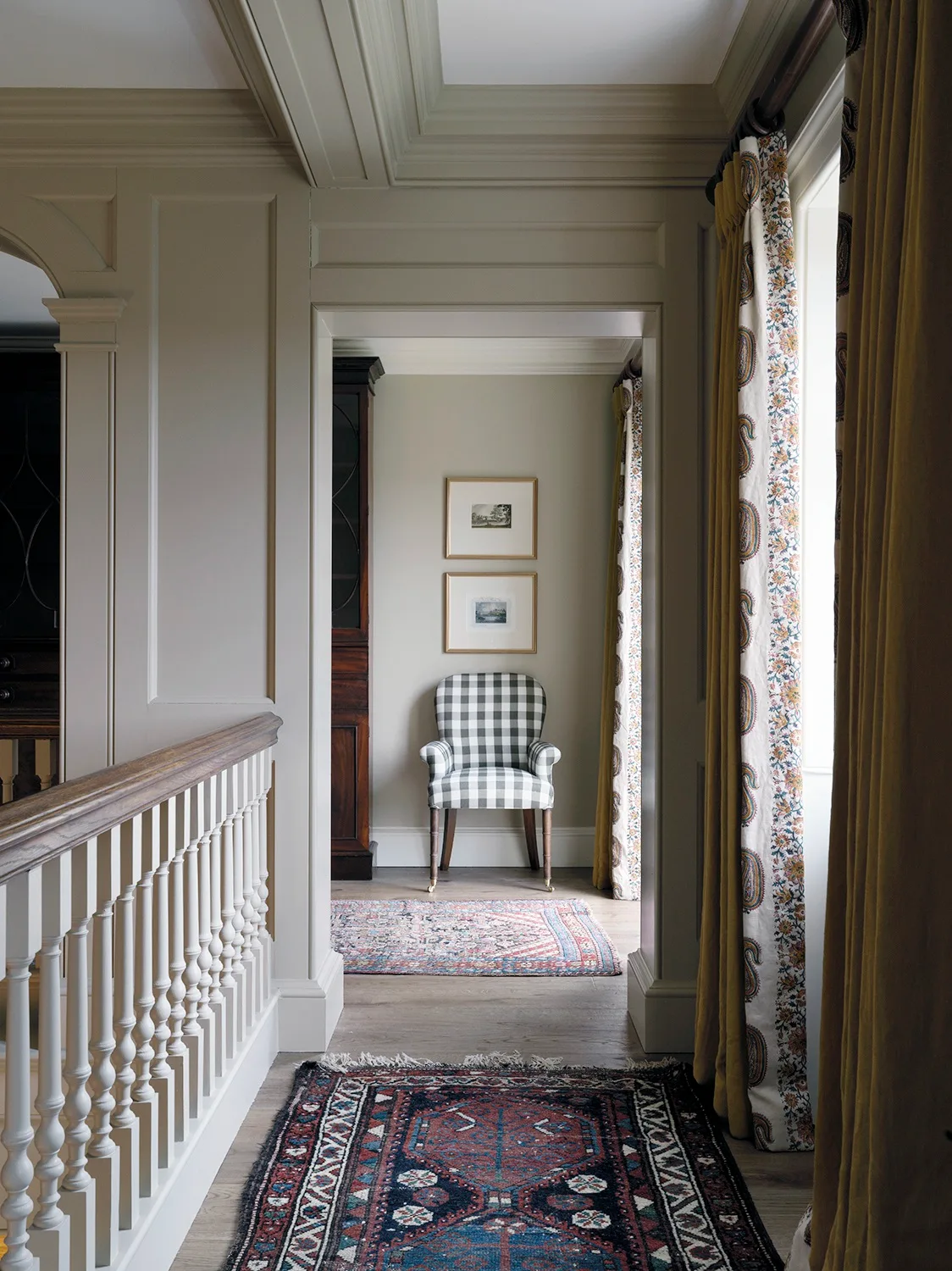 A liminal hallway on the second floor of a home, with coffered ceilings, traditional patterned rugs, heavy curtains and a checkered armchair.