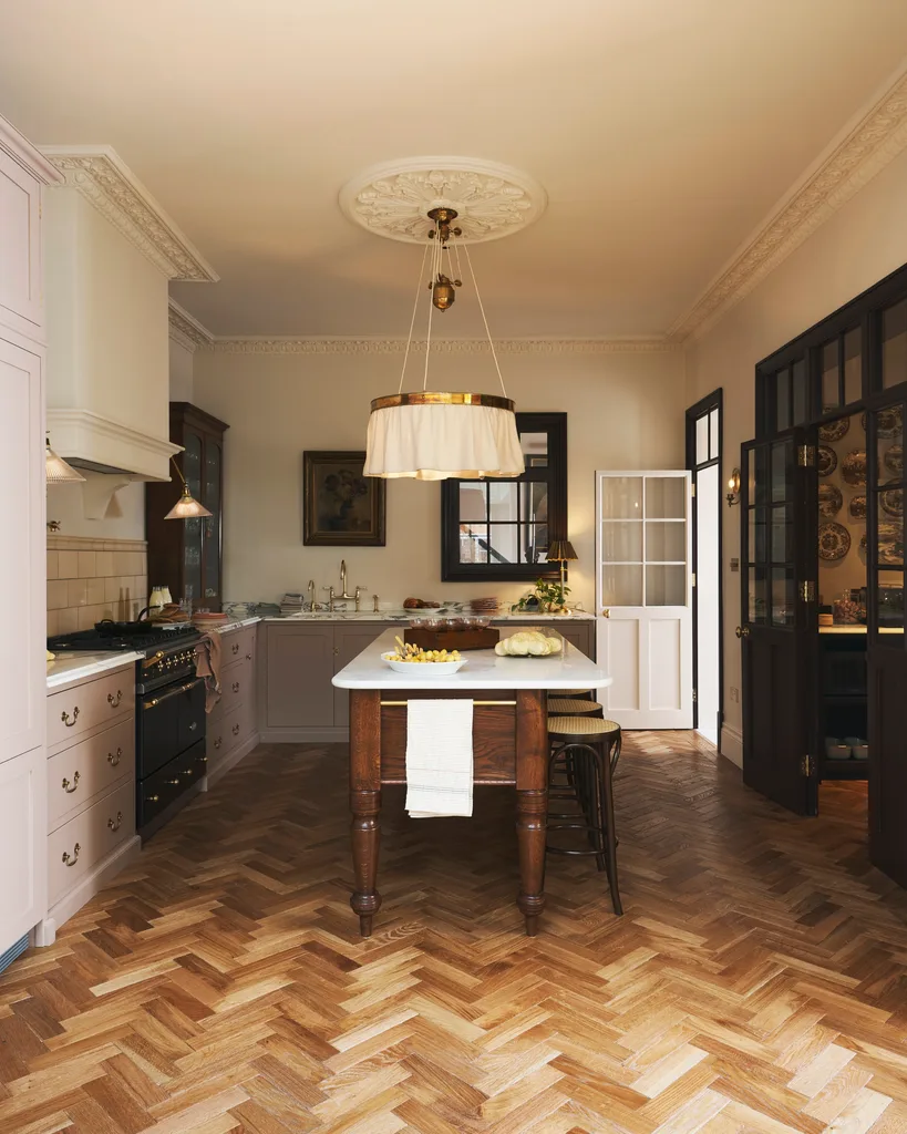A kitchen in an Edwardian home with a splashback made from cream subway tiles, cabinetry with a shaker profile finished in a soft pink shade, a fabric chandelier over a kitchen island with wooden legs and a white marble benchtop. The chandelier is hanging from a ceiling rose.