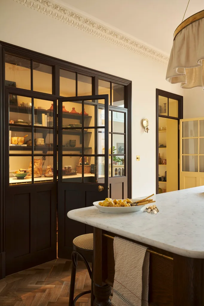 A kitchen in an Edwardian home with a fabric chandelier over a kitchen island with wooden legs and a white marble benchtop. A pantry that is the size of a large walk-in cupboard fits into one wall, with doors that have panels of glass to make the space feel more expansive.