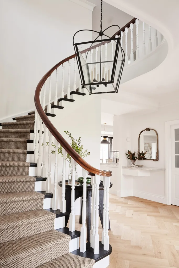 A grand and curving staircase with wooden handrail, white balustrades and a jute stair runner. A chandelier hangs in the void, drawing the eye upwards in this liminal space.