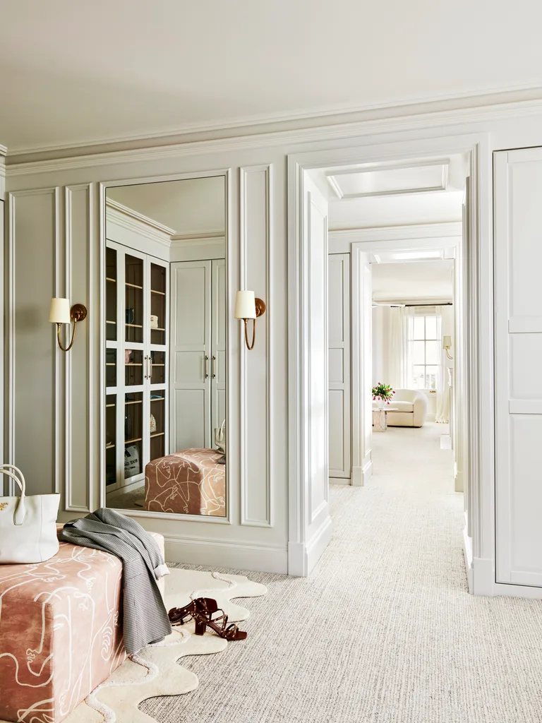 A walk-in wardrobe with white walls that have decorative mouldings, two light sconces with white lampshades, an ottoman in a blush shade with a hangbag on top. The liminal space leads into the bedroom.