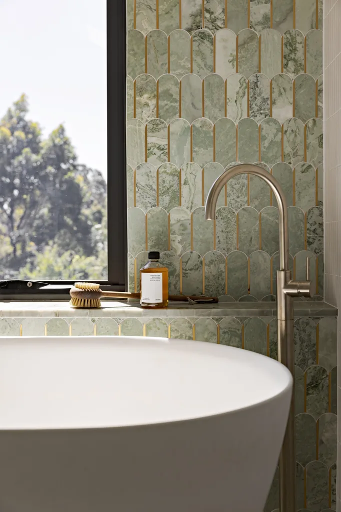 A bathroom with wall tiles in an arch shape, in a sage green colour. A window looks on to vistas of bushland while a curved, freestanding bath is beside Brushed Nickel tapware.