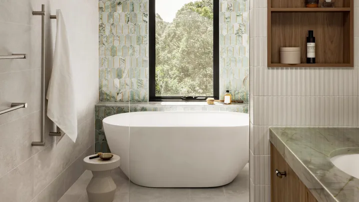 An ensuite bathroom. A vanity is on the right side and in the background is a feature wall with sage green tiles in an arch shape. A window looks on to vistas of bushland behind a curved, freestanding bath.