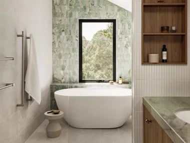 An ensuite bathroom. A vanity is on the right side and in the background is a feature wall with sage green tiles in an arch shape. A window looks on to vistas of bushland behind a curved, freestanding bath.