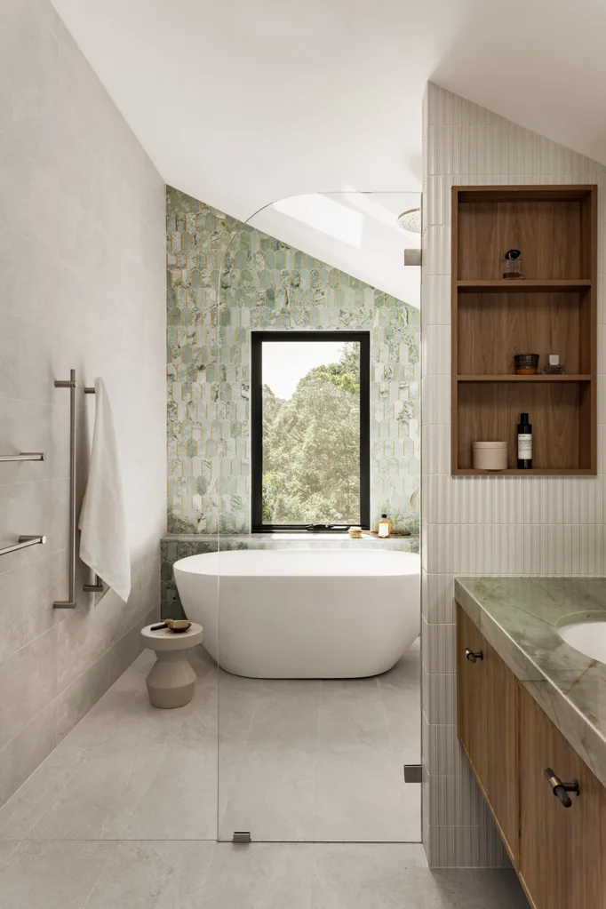 An ensuite bathroom. A vanity is on the right side and in the background is a feature wall with sage green tiles in an arch shape. A window looks on to vistas of bushland behind a curved, freestanding bath.