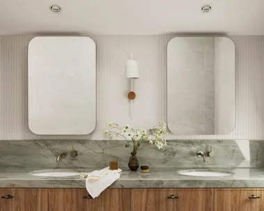 A bathroom vanity with timber cupboards, sage green stone benchtop and splashback, two rectangular mirrors and white wall tiles. A skylight is above.