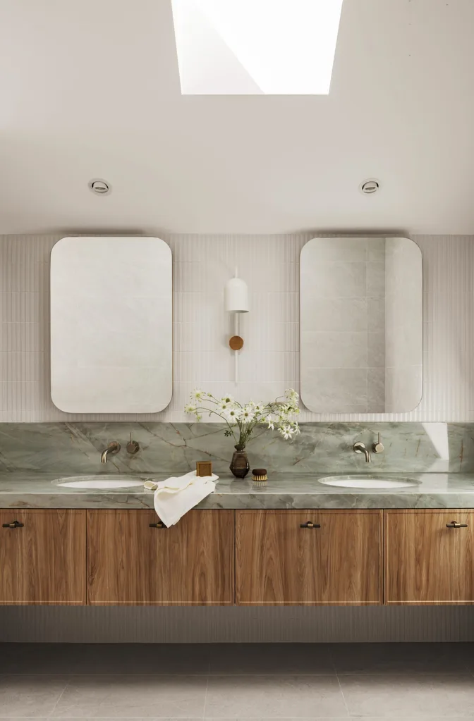A bathroom vanity with timber cupboards, sage green stone benchtop and splashback, two rectangular mirrors and white wall tiles. A skylight is above.