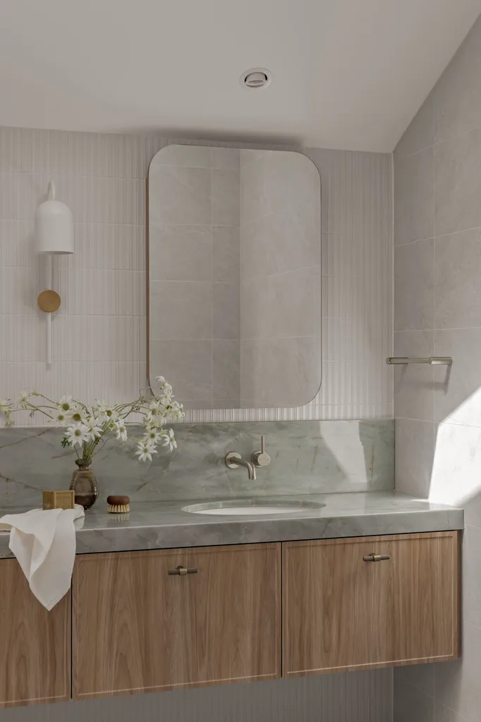 A bathroom vanity with timber cupboards, sage green stone benchtop and splashback, a rectangular mirror and white wall tiles.