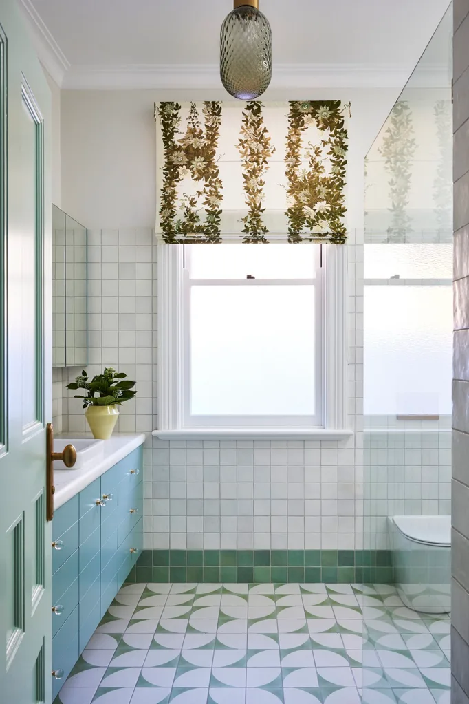 A bathroom in a federation house with tessellated tiles on the floor, teal joinery, white wall tiles and a blind made from a fabric that has a cream background with a green botanical design on it.