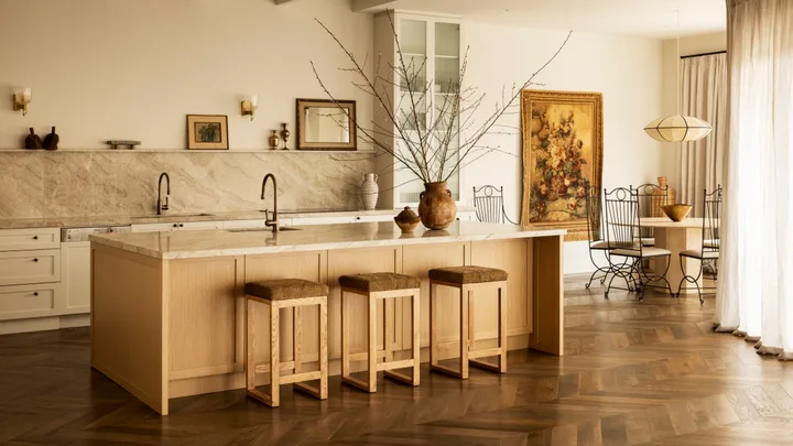 A warm-toned kitchen with a stone splashback and kitchen island, wood floors and soft curtains.
