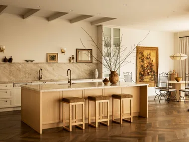 A warm-toned kitchen with a stone splashback and kitchen island, wood floors and soft curtains.
