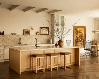 A warm-toned kitchen with a stone splashback and kitchen island, wood floors and soft curtains.