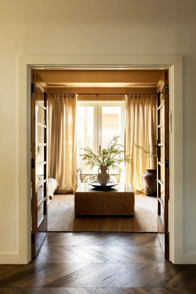 French doors open to a living room with soft yellow curtains, a jute rug and wicker ottoman.