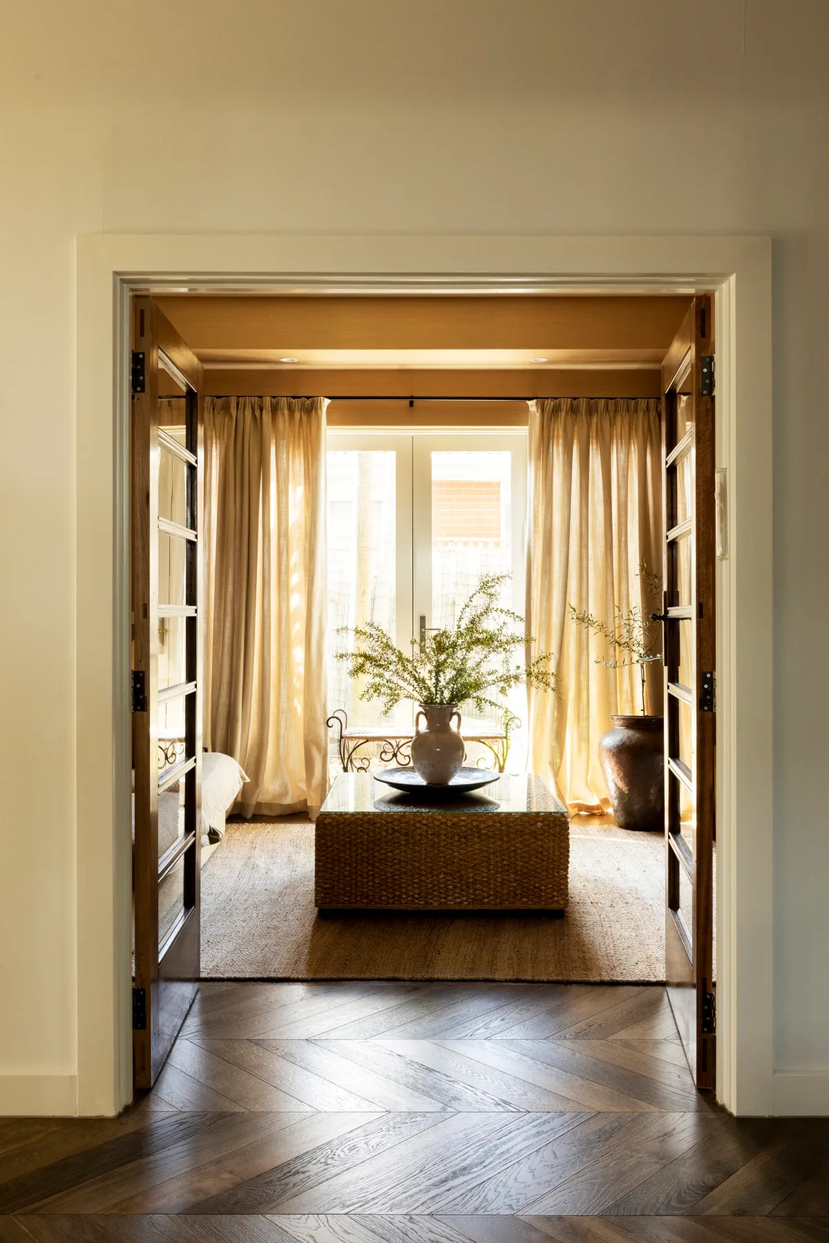 French doors open to a living room with soft yellow curtains, a jute rug and wicker ottoman. 