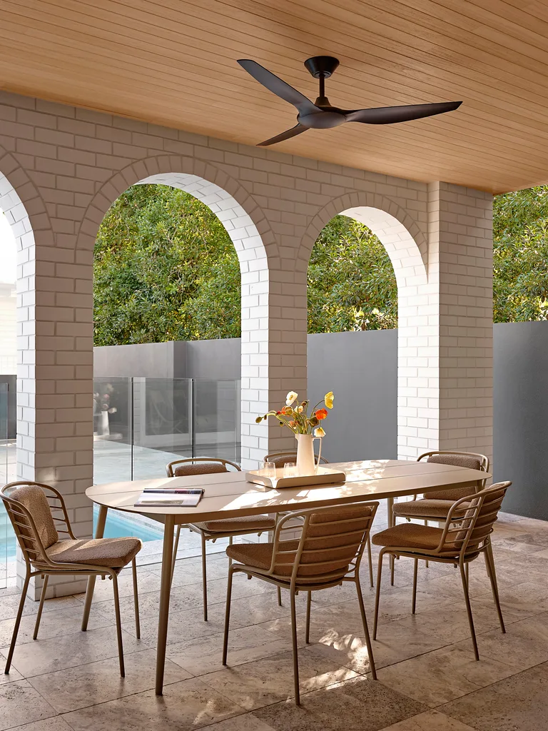 An alfresco dining area. The ceiling is timber, with a black fan attached, while the walls are white brick and curve into three large arches, in a style reminiscent of Spanish mission architecture. The alfresco dining set features a timber table and metal chairs with cushions. A pool is visible in the background. This was all part of an extension and renovation that lasted 13 months.