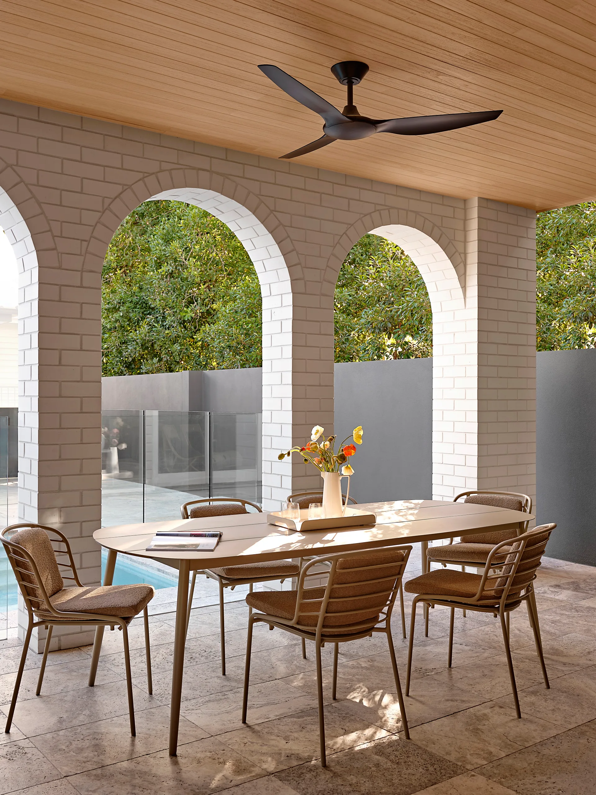 An alfresco dining area. The ceiling is timber, with a black fan attached, while the walls are white brick and curve into three large arches, in a style reminiscent of Spanish mission architecture. The alfresco dining set features a timber table and metal chairs with cushions. A pool is visible in the background. This was all part of an extension and renovation that lasted 13 months.