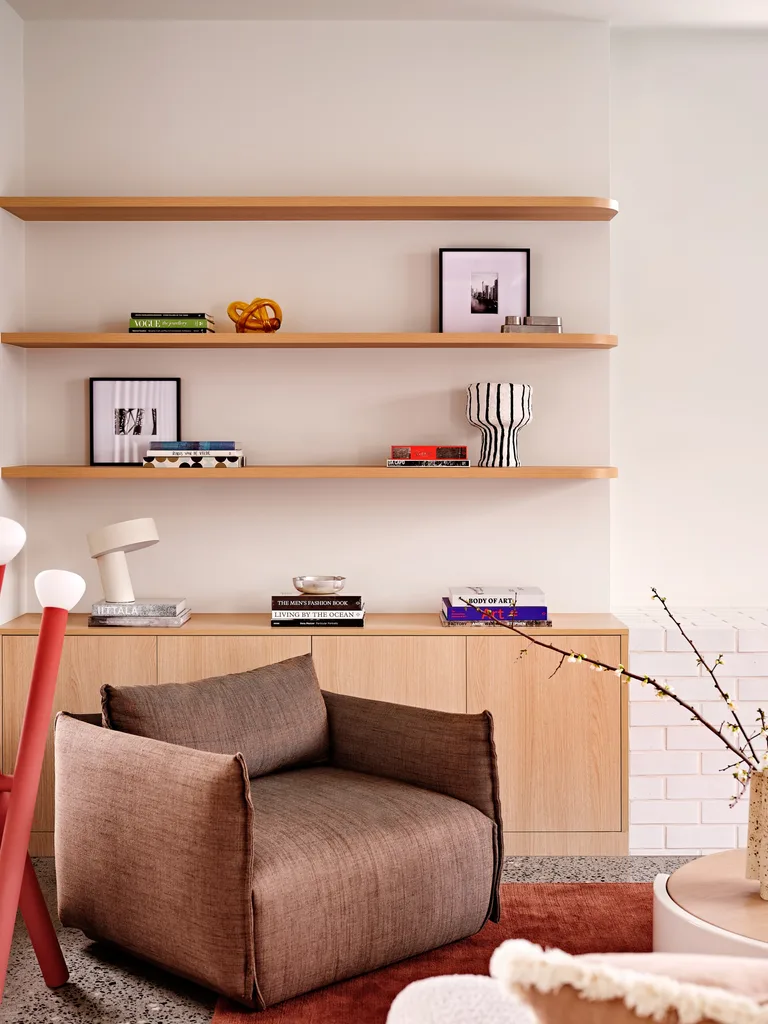A book nook in the corner of a living room. Timber open shelves go across a white wall, above timber cabinetry. An armchair is positioned in front of these shelves, partially on top of the corner of a red rug.