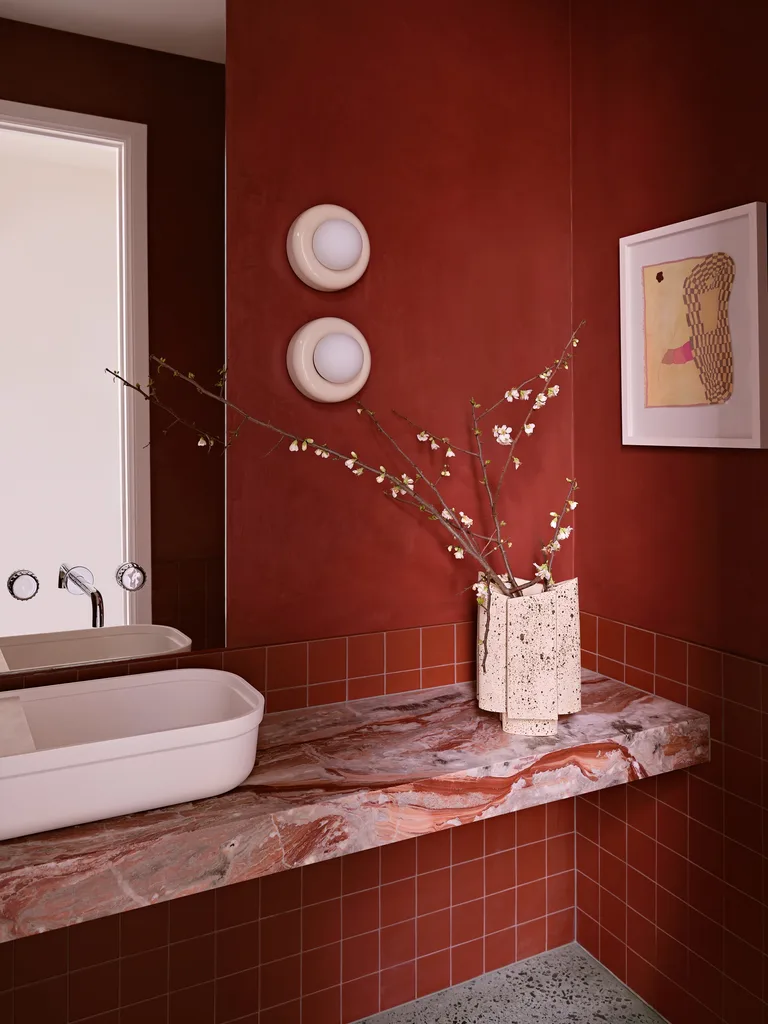 A bathroom vanity with deep, red tones. The walls are finished in a deep red paint, covering two thirds of the height of the wall. Underneath, on the bottom third, are red square tiles. A slab of marble creates a vanity benchtop and also features red tones. The above-counter basin is white. This was all part of an extension and renovation that lasted 13 months.
