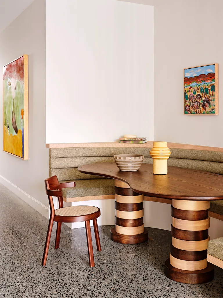 A casual dining nook, with banquette seating that curves around a table in a U-shape. The dining table has an asymmetrical tabletop and a mix of dark and light timber in stripes down the two thick legs. A dining chair is positioned beside with two paintings on the walls.