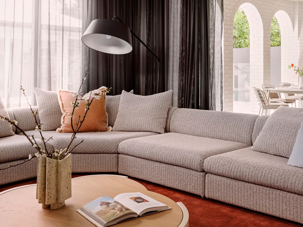 A close up of a grey sofa, that curves around the room in an L shape. Timber coffee tables are in the foreground, on top of a bright red rug. An alfresco dining area is visible in the background. This was all part of an extension and renovation that lasted 13 months.