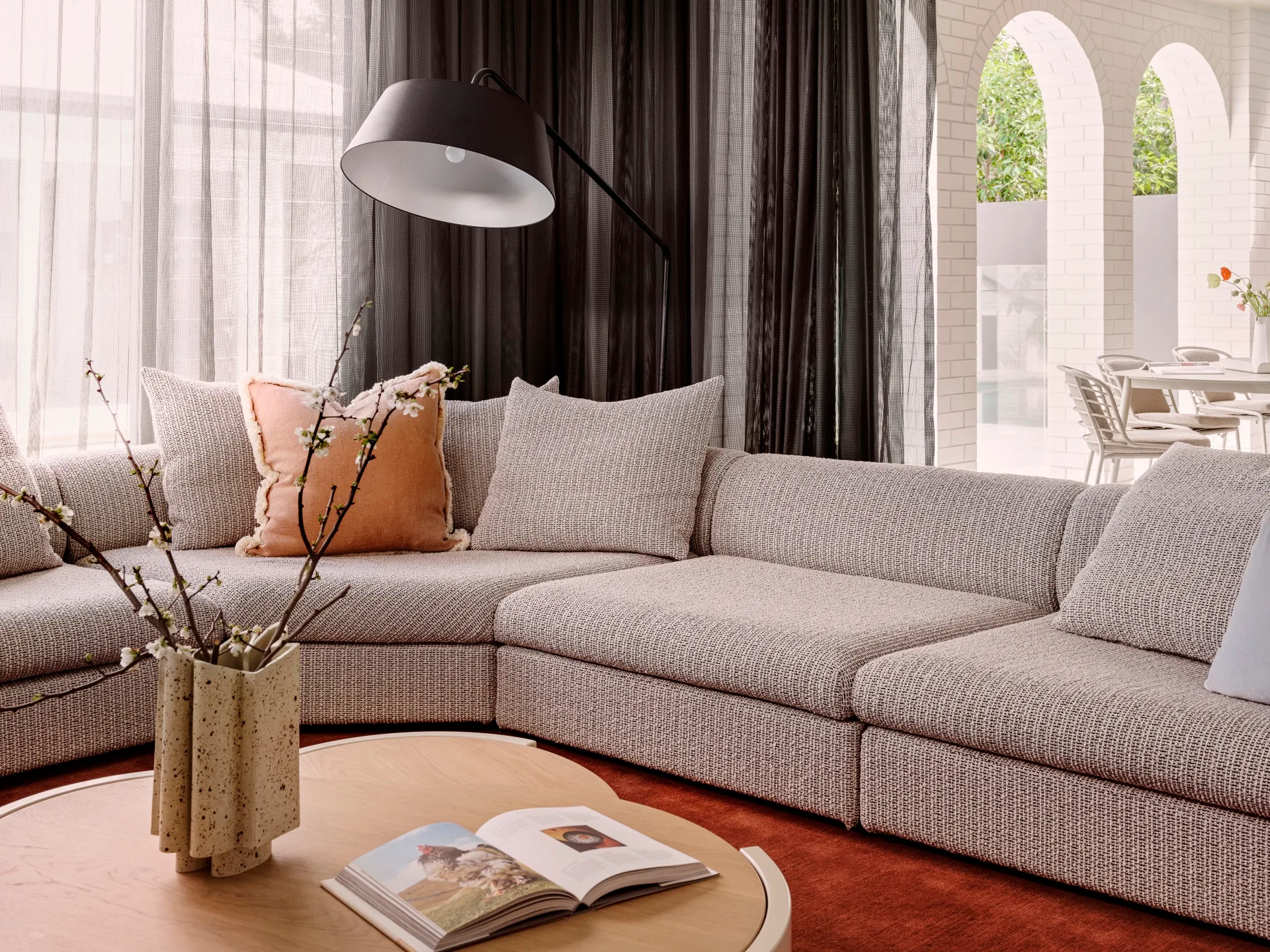 A close up of a grey sofa, that curves around the room in an L shape. Timber coffee tables are in the foreground, on top of a bright red rug. An alfresco dining area is visible in the background. This was all part of an extension and renovation that lasted 13 months.