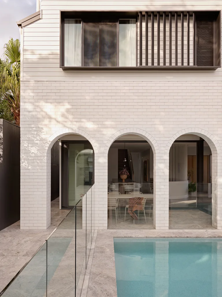 A white brick exterior of a home. The pool, with a glass fence, is in the foreground. Beyond this, arches lead to an alfresco dining area. This was all part of an extension and renovation that lasted 13 months.