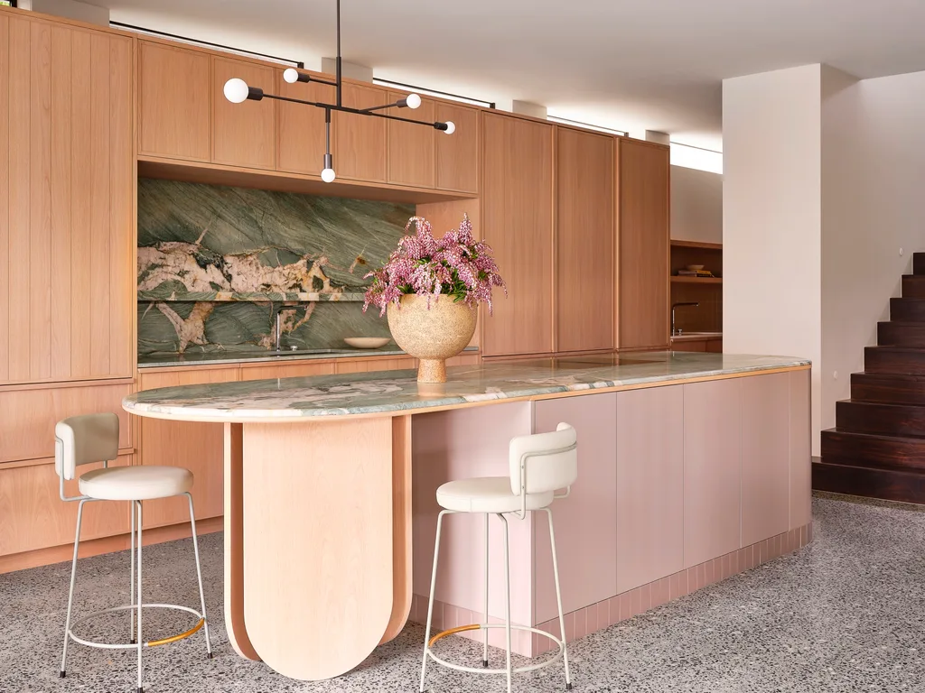 An open plan kitchen with timber joinery, green marble benchtops and splashbacks, and a kitchen island that has a pink profile. Two white bar stools are positioned at one end of the island, which has a benchtop overhang, allowing for casual seating. This was all part of an extension and renovation that lasted 13 months.